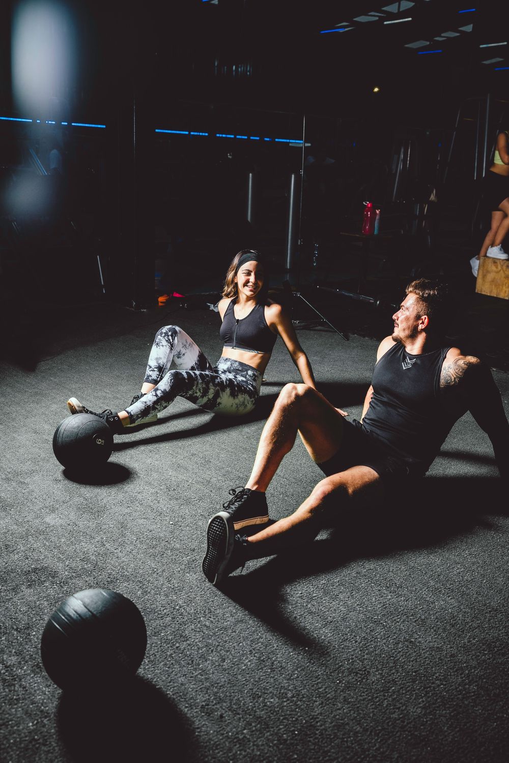 Woman and man sitting on a dark gym floor between sets, smiling, with slam balls nearby